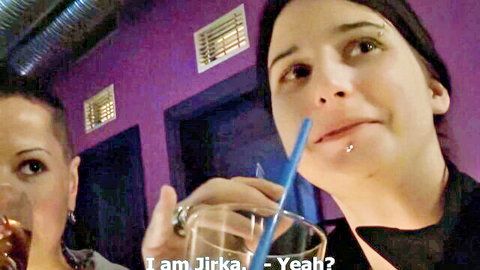 Two pierced Czech women sipping drinks through blue straw in dim purple club bar