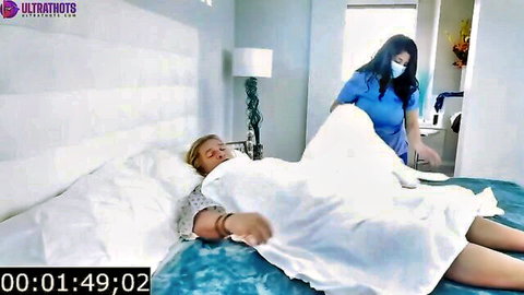 Young Asian nurse in blue scrubs teasingly pulls white sheet off sleeping older blond man's thigh in bed