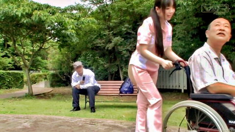 Young Japanese nurse in pink uniform pushes smiling old man in wheelchair through public park outdoors