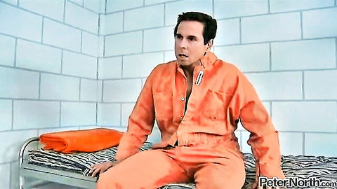 Peter North in orange prison jumpsuit unzipped exposing chest sitting on bunk in jail cell