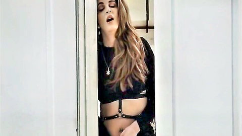 Seductive busty blonde milf in black harness leaning against doorframe