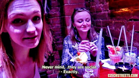 Two busty women in plaid shirts chatting over colorful cocktails in dim bar showing cleavage