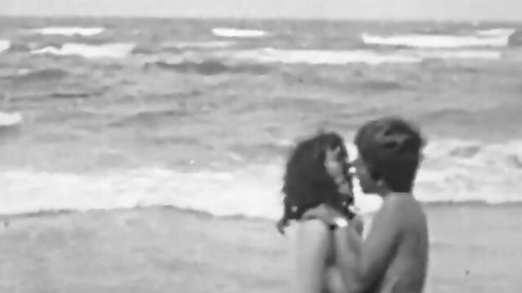 Vintage black and white nude couple intimately embracing on nudist beach with ocean waves crashing behind.