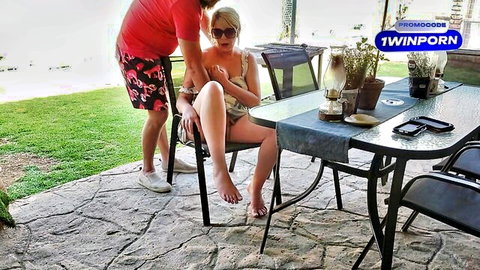Blonde babe in sunglasses spreads legs wide outdoors on patio as guy in swim trunks stands behind her