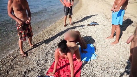 Nude woman bent over on public beach, ass exposed with cum, surrounded by naked men