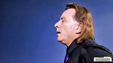 Older brunette man passionately singing in profile wearing black suit purple tie on stage