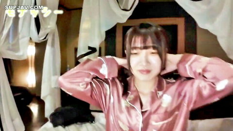 Sexy Asian woman in pink satin pajamas raises arms showing deep cleavage on bed