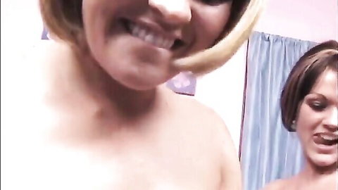 Blonde bob-haired woman smiling wide-mouthed beside brunette in pink bedroom POV