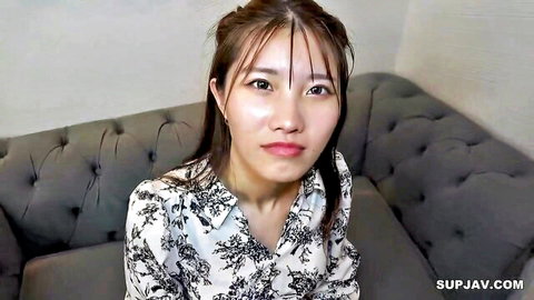 Cute East Asian girl in floral blouse sitting on gray couch with sultry gaze