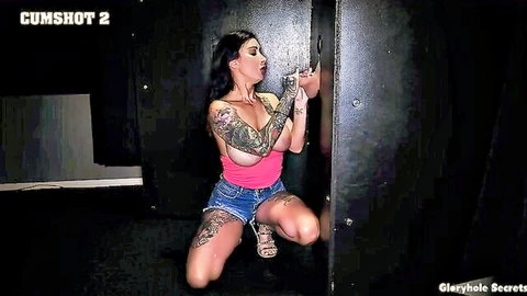 Tattooed brunette kneeling in pink top and denim shorts at gloryhole