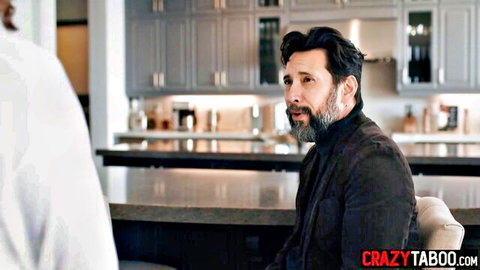 Bearded mature man in dark suit sits at kitchen counter talking to younger man in white shirt