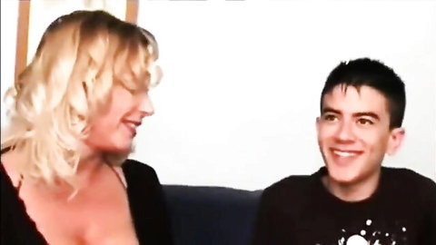 Blonde MILF smiles at teen boy, showing deep cleavage in low-cut top