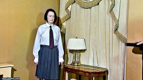 Mature schoolgirl in white shirt, purple tie, and gray pleated skirt stands teasingly in bedroom