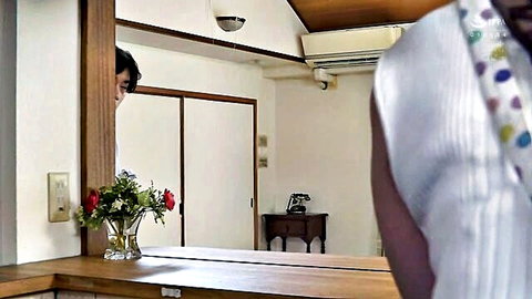 Japanese woman in polka dot top stands at vanity mirror as man peeks from behind in taboo scene