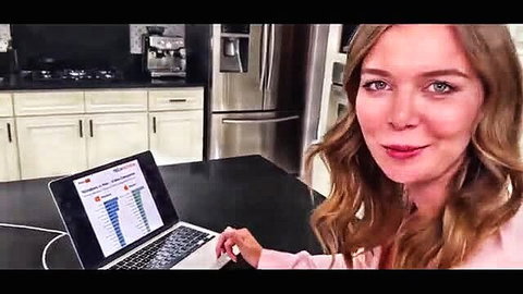 Blonde MILF in kitchen smiles at camera showing laptop graph post-game stress relief