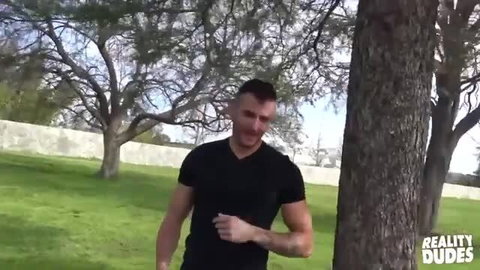 Muscular hunk with buzzcut leaning against tree outdoors, Reality Dudes