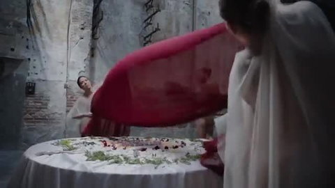 Two pale women in white dresses swirling red fabric over fruit table in erotic scene