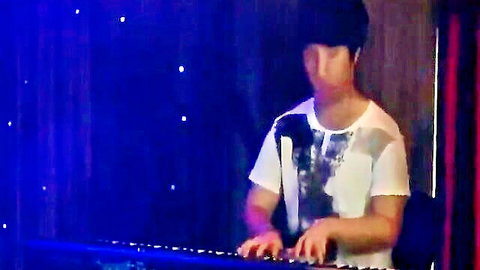 Asian man playing piano on stage in white graphic tee, dim blue lighting