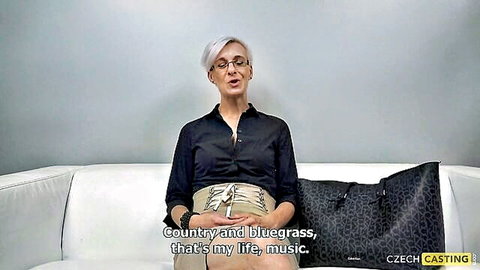 Mature blonde with glasses chats on Czech casting couch in black shirt and beige pants