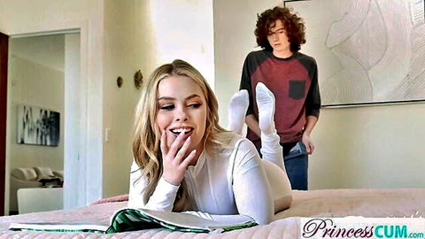Blonde in white bodysuit and socks lying on bed reading, guy watching from behind