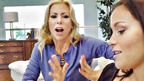 Blonde MILF gesturing intensely at surprised brunette babe on couch