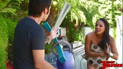 Hot ebony babe in polka dot sarong laughs with guy holding giant dildo poolside