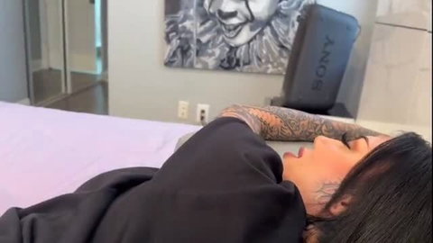 Tattooed Lacey Jayne lying on bed in black hoodie, open mouth tease