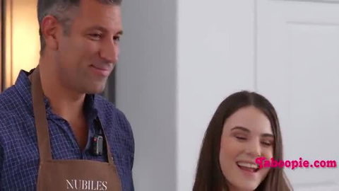 Stepdad in apron smiles at smiling young brunette stepdaughter in kitchen
