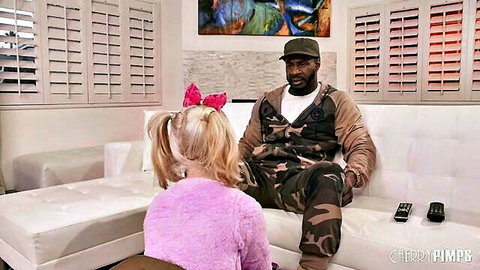 Blonde girl with pigtails in pink sweater sits with black man in camo on couch