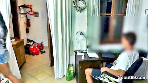 German teen in short denim shorts stands near blurred man at desk in amateur home setup
