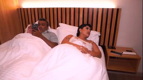 Man scrolls phone in bed next to busty Latina stepmom in lingerie under covers