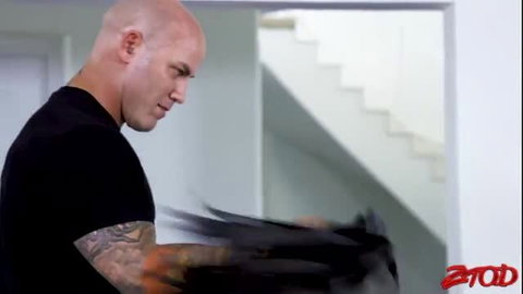Bald tattooed muscular man in black shirt holding dark clothing in Zero Tolerance scene