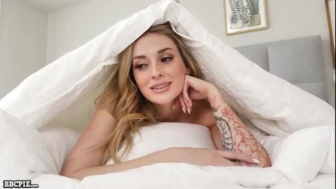 Blonde tattooed babe peeking seductively from white sheets in bed