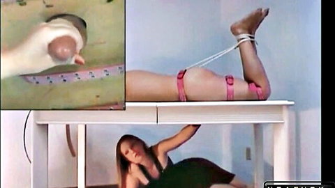 Skinny brunette with small tits has legs bound spread wide exposing bare pussy under glory hole table grabbing thick cock