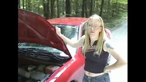Blonde woman in glasses stands by open red car hood in forest