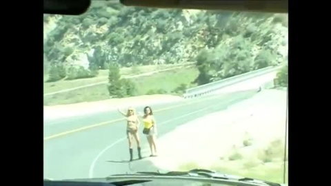 Two women hitchhiking topless on a mountain road in daylight