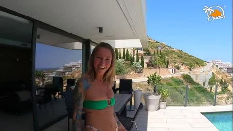 Blonde tattooed woman in green bikini smiling on sunny poolside balcony