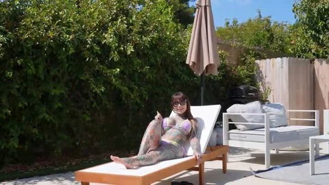 Tattooed woman in micro bikini posing on poolside lounge chair