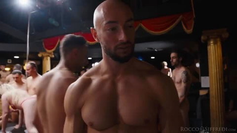 Bald muscular man with beard in nightclub, shirtless, surrounded by male performers