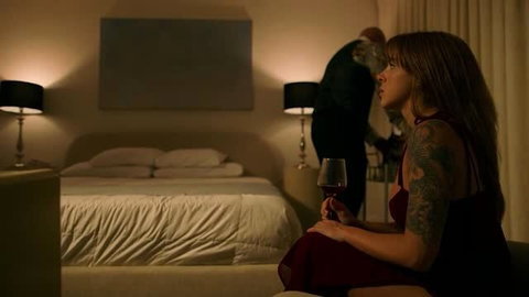 Tattooed woman in red dress with wine glass watches bald man in suit