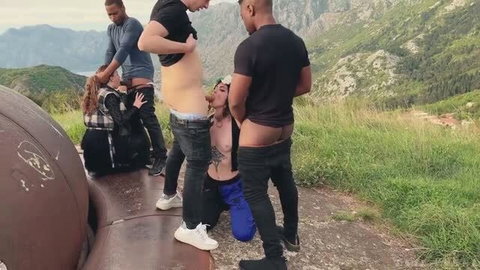 Outdoor group sex scene with tattooed woman on knees servicing multiple men