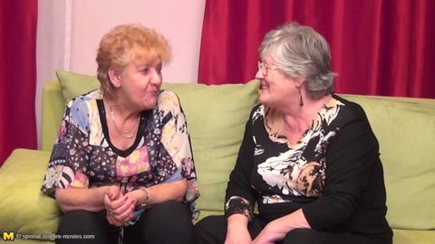Two mature lesbian grandmas on couch smiling at each other