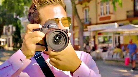 Blonde man in pink shirt sunglasses holding Sony camera to face photographing sunny street cafe scene