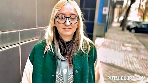 Blonde teen with glasses in green varsity jacket posing sexy on urban street