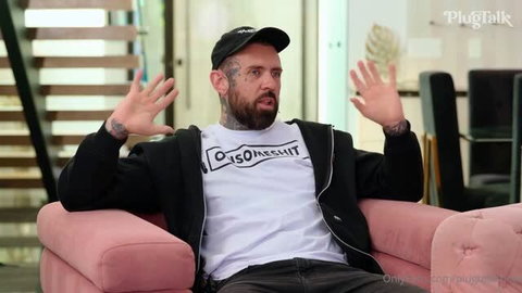 Lena The Plug in black cap and white t-shirt on pink couch, arms raised