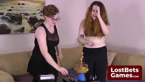 Two curvy women play strip game, one in sheer black dress, one topless