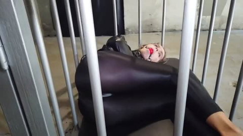 Woman in shiny black catsuit and red ball gag locked in metal cage