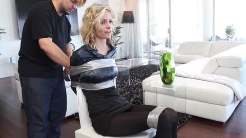 Blonde woman in black dress duct taped to chair by man in t-shirt