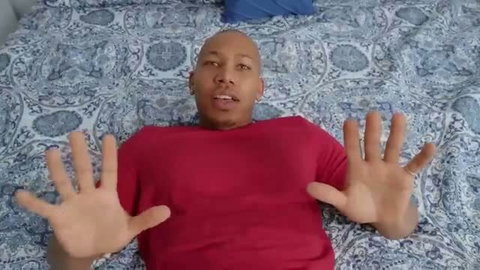 Bald Black man in red shirt lying on bed with hands up
