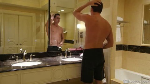 Shirtless muscular man in black shorts checking mirror reflection in hotel bathroom
